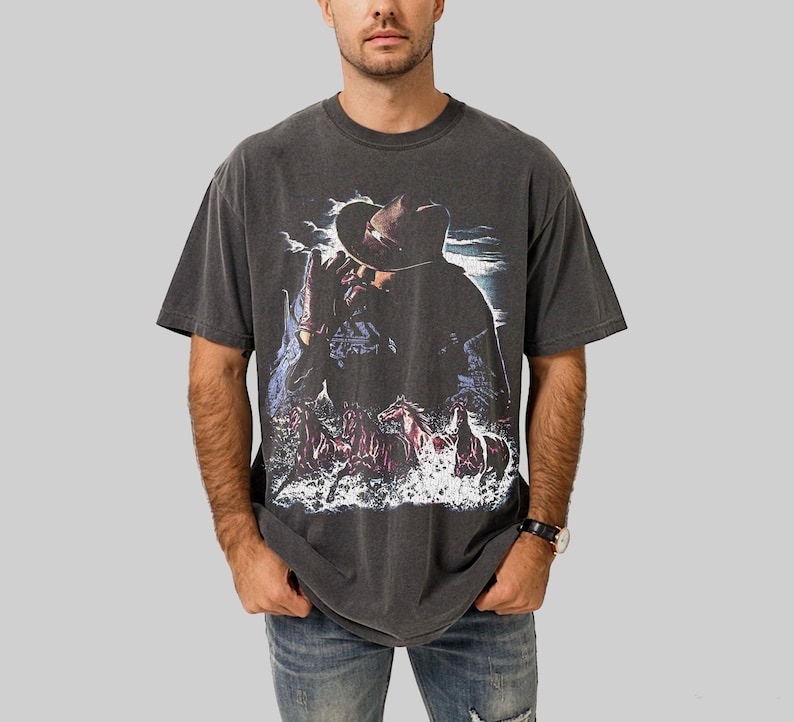 Puede incluir: Camiseta gris oscuro con un gr&aacute;fico de vaquero y caballos en el agua. El dise&ntilde;o incluye un vaquero con sombrero, con un aspecto vintage y desgastado. La camiseta es de cuello redondo y manga corta.