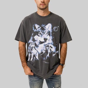 May include: Dark gray t-shirt featuring a detailed wolf graphic with a full moon and starry sky. The design includes multiple wolves and mountain silhouettes. The shirt has a vintage, washed look.