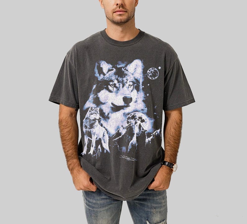 May include: Dark gray t-shirt featuring a detailed wolf graphic with a full moon and starry sky. The design includes multiple wolves and mountain silhouettes. The shirt has a vintage, washed look.