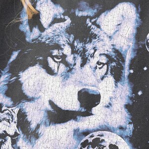 May include: Dark gray t-shirt featuring a detailed wolf graphic with a celestial theme. The design includes a large wolf face, smaller wolf, and planetary bodies in shades of blue and white.