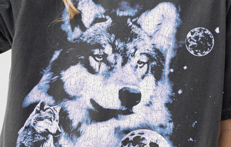 May include: Dark gray t-shirt featuring a detailed wolf graphic with a celestial theme. The design includes a large wolf face, smaller wolf, and planetary bodies in shades of blue and white.