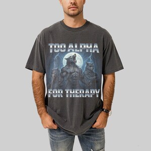May include: Dark gray t-shirt featuring a graphic of three werewolves howling at the moon, with lightning bolts in the background. The text "TOO ALPHA FOR THERAPY" is printed above and below the graphic in a silver, metallic font.