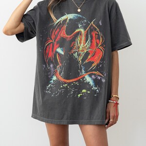 May include: Dark grey t-shirt with a striking graphic of a red and orange dragon with large wings. The dragon is set against a dark background and perched on a rocky structure. The shirt has short sleeves and a relaxed fit.