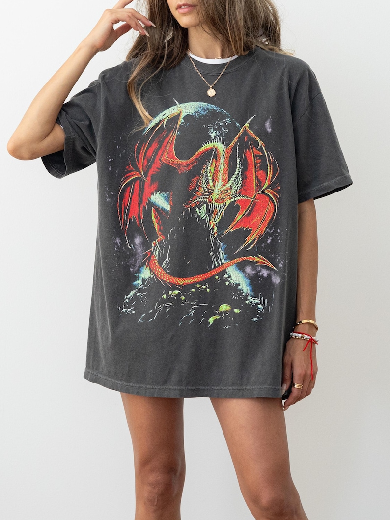 May include: Dark grey t-shirt with a striking graphic of a red and orange dragon with large wings. The dragon is set against a dark background and perched on a rocky structure. The shirt has short sleeves and a relaxed fit.