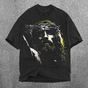 May include: Black t-shirt featuring a distressed graphic of a person wearing a crown of thorns. The graphic has yellow and white accents. The shirt has short sleeves and a crew neck.