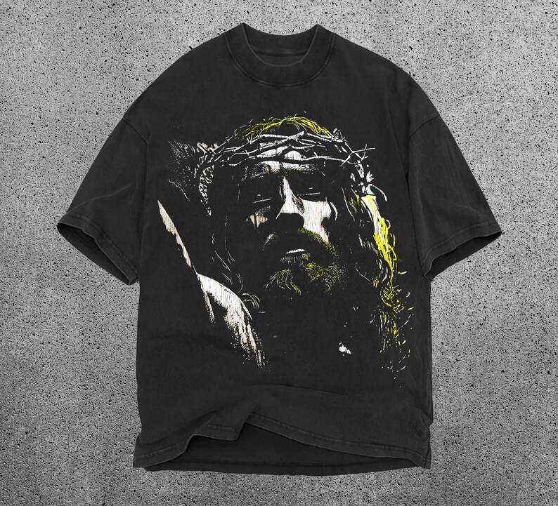 May include: Black t-shirt featuring a distressed graphic of a person wearing a crown of thorns. The graphic has yellow and white accents. The shirt has short sleeves and a crew neck.