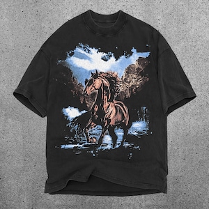 May include: Black t-shirt featuring a brown horse graphic. The horse is depicted in a running pose against a backdrop of a blue sky and trees. The shirt has a crew neck and short sleeves.