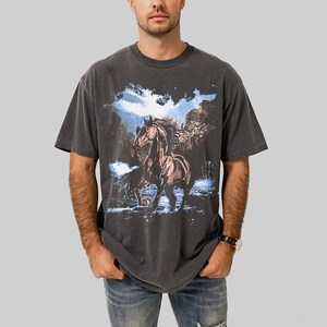 May include: Dark gray t-shirt featuring a brown horse in a mountain landscape with blue sky and water. The graphic tee has a vintage, washed look and a classic crew neck.