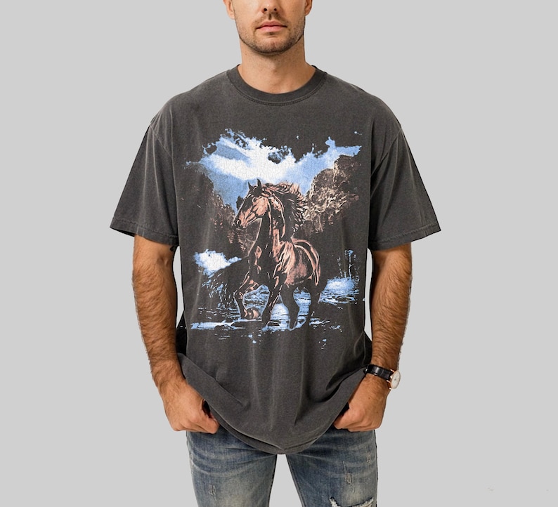May include: Dark gray t-shirt featuring a brown horse in a mountain landscape with blue sky and water. The graphic tee has a vintage, washed look and a classic crew neck.