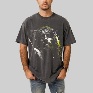 May include: Dark gray t-shirt featuring a graphic of a person with a crown of thorns. The image has yellow and green accents. The shirt is a short-sleeved, crew neck style.