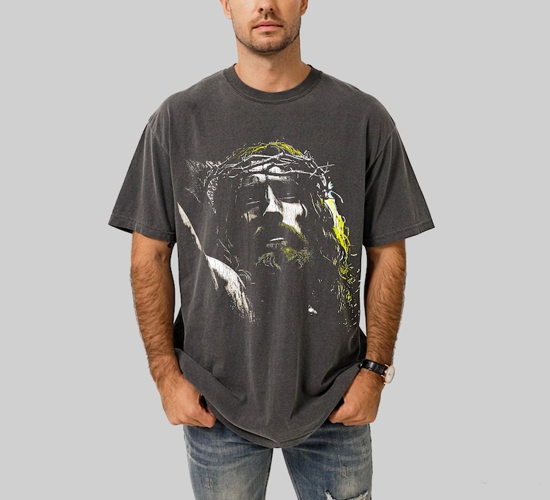 May include: Dark gray t-shirt featuring a graphic of a person with a crown of thorns. The image has yellow and green accents. The shirt is a short-sleeved, crew neck style.