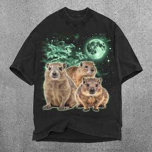 May include: Black t-shirt featuring a graphic of three groundhogs under a glowing green moon and starry sky. The groundhogs are brown and tan with a green outline. The background has green clouds.