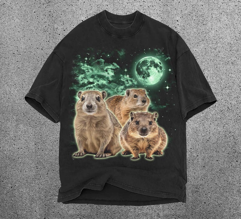 May include: Black t-shirt featuring a graphic of three groundhogs under a glowing green moon and starry sky. The groundhogs are brown and tan with a green outline. The background has green clouds.