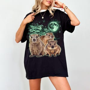 May include: Black t-shirt featuring a graphic of three brown rock hyraxes under a green moon and starry sky. The shirt has a relaxed fit and a white collar peeking out from the neckline.