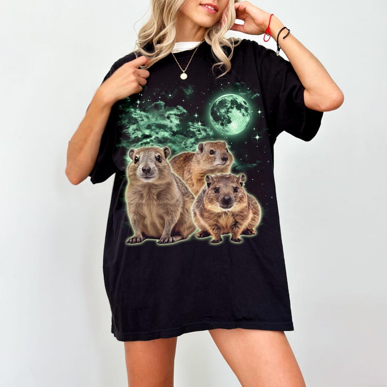 May include: Black t-shirt featuring a graphic of three brown rock hyraxes under a green moon and starry sky. The shirt has a relaxed fit and a white collar peeking out from the neckline.