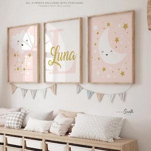 Könnte beinhalten: Drei rosa und goldene Kinderzimmerdrucke mit einer Wolke, einem Buchstaben L mit dem Namen Luna und einem Halbmond. Die Drucke sind gerahmt und an einer weißen Wand über einer Holzaufbewahrungsbank mit weißen Behältern aufgehängt.