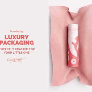 May include: A cylindrical product container with a white and coral design, resting on a pink fabric. The container has text that reads "Thank you for choosing" and "Luxury Packaging" in bold letters. The image also includes the text "Perfectly crafted for your little one."