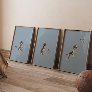 May include: Three framed prints featuring watercolor illustrations of dogs and birds on a muted blue background. The dogs are brown and white, and the birds are depicted in flight. The frames are a light wood color.