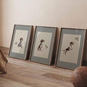 Op de afbeelding: Drie ingelijste kunstdrukken van honden in aquarel. Elke print toont een andere hondenpose met een eend. De frames hebben een houten afwerking en een gestreepte rand in blauw en wit.