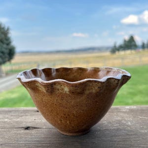 May include: A handmade ceramic bowl with a ruffled rim. The bowl is a warm brown color with a speckled texture and a white rim. The bowl is sitting on a wooden surface, with a blurred outdoor background.
