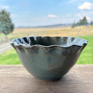 May include: A handmade, teal-colored ceramic bowl with a ruffled rim. The bowl has a speckled texture and sits on a wooden surface. The background features a blurred landscape with green grass and a blue sky.