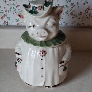 May include: A ceramic pig cookie jar featuring a cream-colored body with a green ruffled collar. The pig wears a matching hat adorned with painted strawberries and leaves. The front of the jar displays a small floral detail and three brown dots.