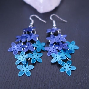 May include: A pair of dangling earrings featuring a cascade of blue flower charms. The earrings showcase a gradient of blue hues, transitioning from deep navy to light turquoise. The floral design adds a delicate and feminine touch.