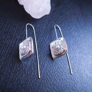 May include: A pair of silver-toned drop earrings. Each earring features a square-shaped pendant with a textured design and a small, round, pearl-like bead in the center. The earrings have long, thin, silver wires with gold-colored tips.