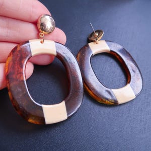 May include: A pair of oval-shaped earrings with a tortoiseshell pattern and a cream-colored accent. The earrings have a gold-colored stud and a post for pierced ears. The design is a mix of brown, amber, and cream colors.
