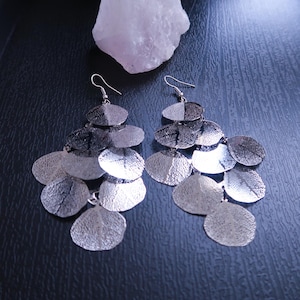 May include: Silver-toned dangle earrings with a leaf-like design. Each earring features multiple overlapping, textured, circular shapes, creating a layered effect. The top portion of each leaf is a darker silver, while the bottom is a lighter silver.