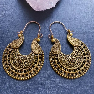 May include: A pair of antique gold-toned metal hoop earrings featuring an intricate filigree pattern with swirling scrolls, floral motifs, and a beaded outer edge. The earrings have a curved wire hook closure and a crescent-shaped design.
