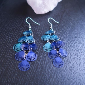 May include: A pair of dangling earrings featuring a cascade of blue seashell-shaped charms. The earrings have a gradient of blue hues, from light turquoise to deep navy. The hooks are silver-colored.