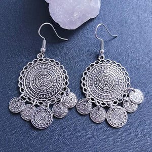 May include: A pair of silver-colored dangle earrings. Each earring features a large, circular pendant with intricate patterns and smaller, coin-shaped charms hanging below. The earrings have a hook closure.