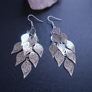 May include: Silver-toned leaf earrings. Each earring features multiple leaf-shaped pendants with intricate vein detailing, suspended from a simple hook. The leaves are arranged in a cascading design, creating a delicate and natural aesthetic. The earrings are displayed against a dark background.