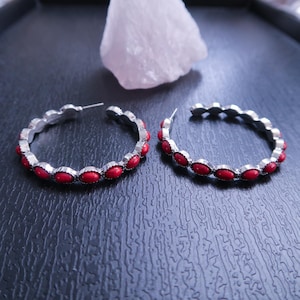 May include: A pair of silver-toned hoop earrings with a row of oval-shaped red stones. The earrings are set against a dark background with a white, irregularly shaped object in the background. The earrings are a jewelry item.