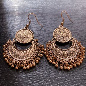May include: A pair of gold-toned dangle earrings with a crescent moon design and a round, ornate pendant. The earrings have a beaded fringe.