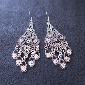 May include: A pair of silver-toned dangle earrings featuring an intricate filigree diamond-shaped top section with floral patterns, connected to multiple dangling teardrop-shaped charms, each containing a small floral motif.