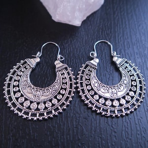 May include: A pair of silver-toned hoop earrings featuring a crescent-shaped design with intricate filigree patterns, small circular cutouts, and a beaded edge. The earrings have a hinged wire closure at the top and a polished metallic finish.