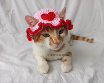 May include: A cat wearing a pink and red crochet hat with a heart design. The hat has a ruffled brim and is placed on the cat's head. The cat has orange and white fur and is lying on a white surface. The text "@FURREVERCROCHET" is visible.