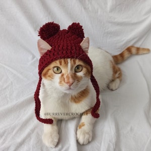 Könnte beinhalten: Eine rot-weiße Katze trägt eine weinrote Häkelmütze mit zwei Bommeln und Ohrenklappen. Die Katze liegt auf einer weißen Oberfläche. Auf der Mütze steht der Text "@FURREVERCROCHET".