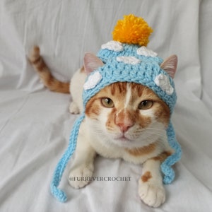May include: A ginger and white cat wearing a light blue crocheted hat with white cloud accents and a yellow pom-pom. The hat has long blue ties. The cat is lying down on a white surface.