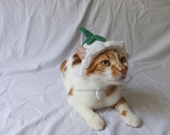 Op de afbeelding: Een wit-oranje kat draagt een handgemaakte gehaakte hoed. De hoed is wit met een groen bladerontwerp. De kat ligt op een wit laken. De tekst "@FURREVERCROCHET" is zichtbaar.