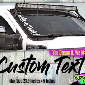 May include: White custom text decal on a white truck windshield. The decal is 23.5 inches by 5 inches. The text reads "Custom Text".