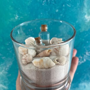 May include: A glass jar filled with white sand and seashells, with two small glass bottles with cork tops.