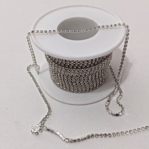 May include: A spool of silver rhinestone chain. The chain is wound around a white plastic spool. A length of the chain extends from the spool, showcasing the sparkling, clear rhinestones. The chain is suitable for jewelry making and craft projects.