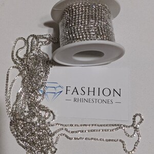 May include: A roll of silver rhinestone chain sits next to a length of the same chain. A white card with the text "FASHION - RHINESTONES" is also in the image. The rhinestones are small and round, and the chain is flexible.