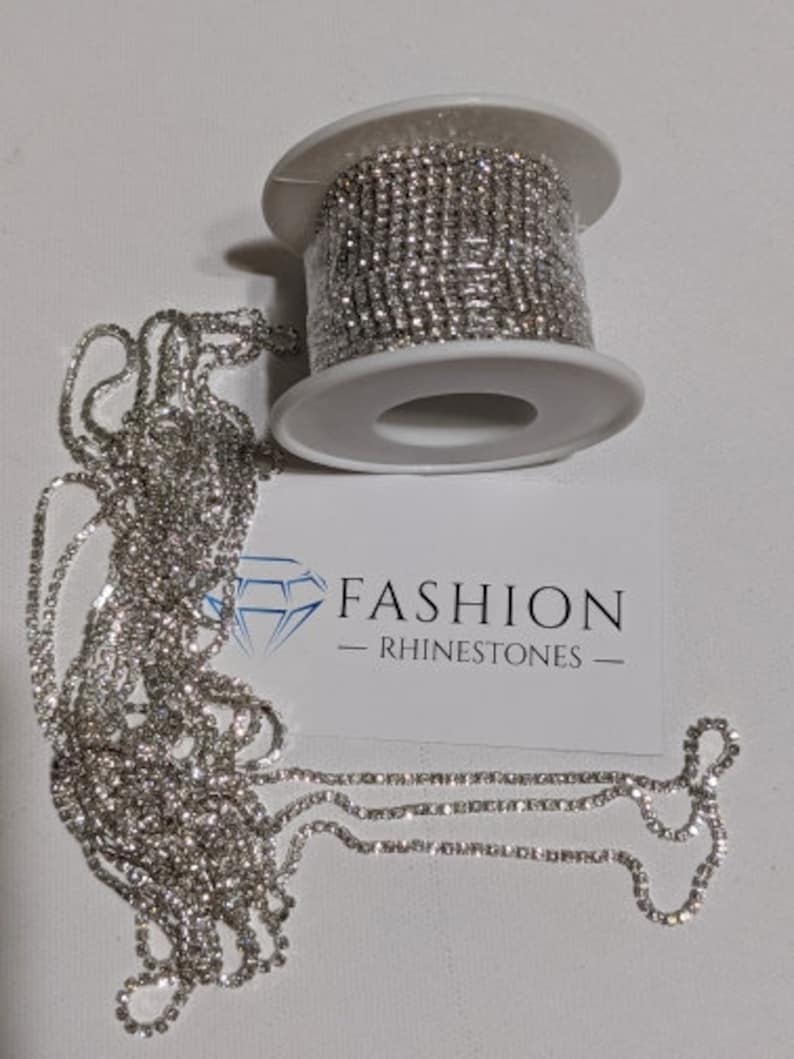May include: A roll of silver rhinestone chain sits next to a length of the same chain. A white card with the text "FASHION - RHINESTONES" is also in the image. The rhinestones are small and round, and the chain is flexible.