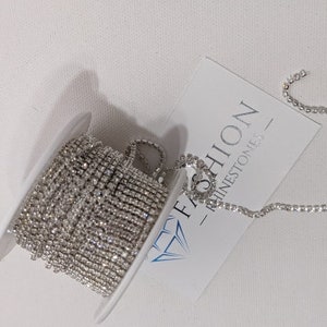 May include: A spool of silver rhinestone chain with a white label that reads "FASHION RHINESTONES". The chain is coiled on a white plastic spool, with a portion of the chain unraveled.