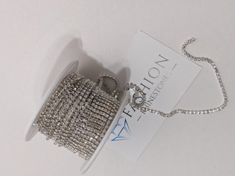 May include: A spool of silver rhinestone chain with a white label that reads "FASHION RHINESTONES". The chain is coiled on a white plastic spool, with a portion of the chain unraveled.