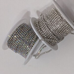 May include: Two spools of rhinestone chain. One spool features clear rhinestones, while the other has iridescent rhinestones. The spools are white and are set against a white background. The rhinestone chain is likely used for jewelry making or embellishments.